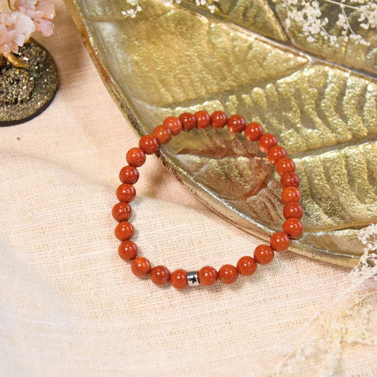 Bracelet en perles de jaspe rouge posé sur un tissu beige texturé, adossé à une coupelle dorée en forme de feuille, entouré d'éléments végétaux et d'un petit arbre en pierres naturelles.