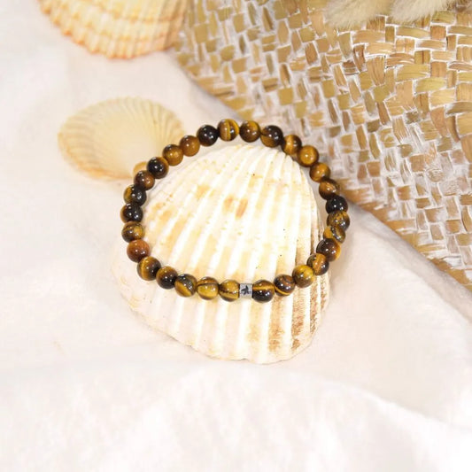 Bracelet en œil de tigre reposant sur un grand coquillage blanc, entouré de tissu beige texturé et d'éléments tressés dans une ambiance douce et solaire.