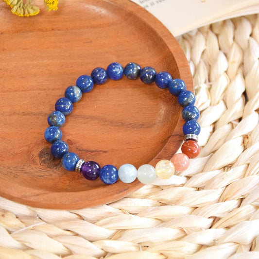 Bracelet en perles de lapis-lazuli et pierres des 7 chakras posé élégamment sur une coupelle en bois brut, elle-même placée sur un set de table en paille tressée. Une touche végétale jaune et un livre ouvert complètent cette scène à l'ambiance bohème et naturelle.