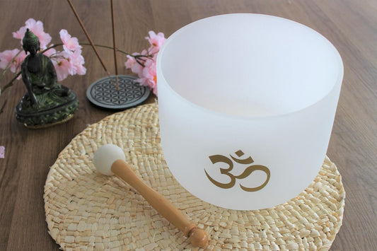 Bol chantant en cristal de quartz blanc Om posé sur un dessous de plat en paille tressée, entouré d'une statuette de Bouddha, d'encens et de fleurs de cerisier.