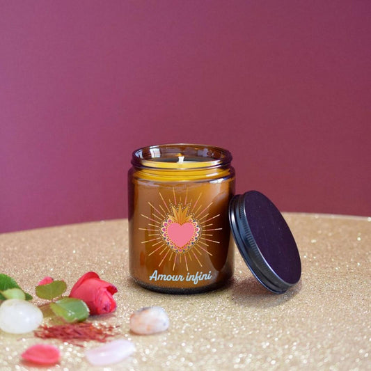 Bougie d'intention Amour infini ouverte sur une surface dorée scintillante, entourée de pétales de rose rouge et de petites pierres naturelles sur fond pourpre chaleureux.