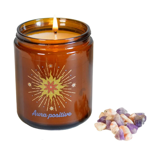 Bougie d'intention en pot de verre ambré allumée avec sa flamme douce, posée à côté d'un petit tas de pierres naturelles brutes d'améthyste et de pierre de soleil sur fond blanc.