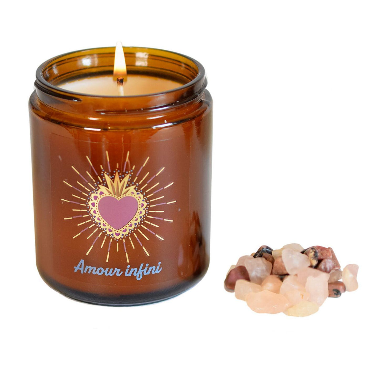 Bougie en verre ambré allumée avec sa flamme douce, posée à côté d'un petit tas de pierres naturelles brutes de quartz rose et de rhodonite sur fond blanc.