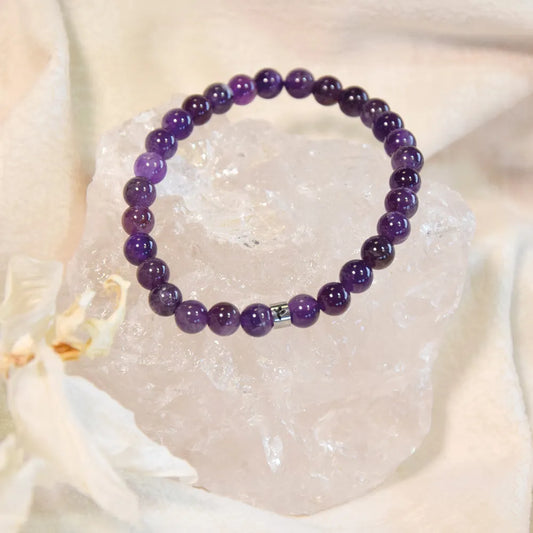 Bracelet de perles rondes en améthyste violette reposant sur un grand cristal de roche brut, avec une perle séparatrice argentée et un pétale de fleur blanche à gauche, sur un tissu beige texturé.
