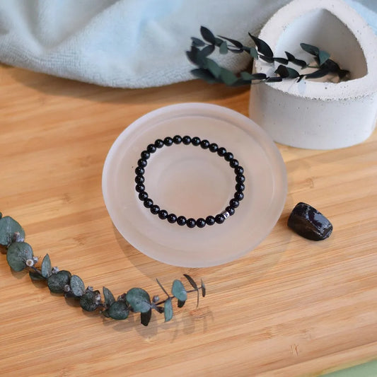 Bracelet en perles de tourmaline noire posé dans une coupelle en sélénite translucide sur un plateau en bois, entouré de branches d'eucalyptus séché et d'une pierre brute.