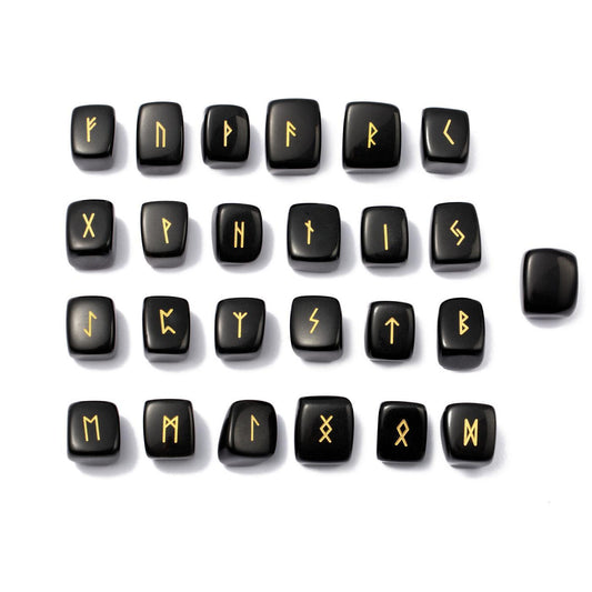 Les 24 runes en obsidienne noire gravées de doré soigneusement alignées en quatre rangées avec la rune vierge posée à droite, présentées à plat sur un fond blanc.