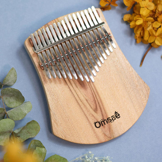 Kalimba en bois de 17 lames posé en biais sur un fond bleu doux, entouré de branches d'eucalyptus et de fleurs séchées jaunes et blanches pour une ambiance poétique et zen.